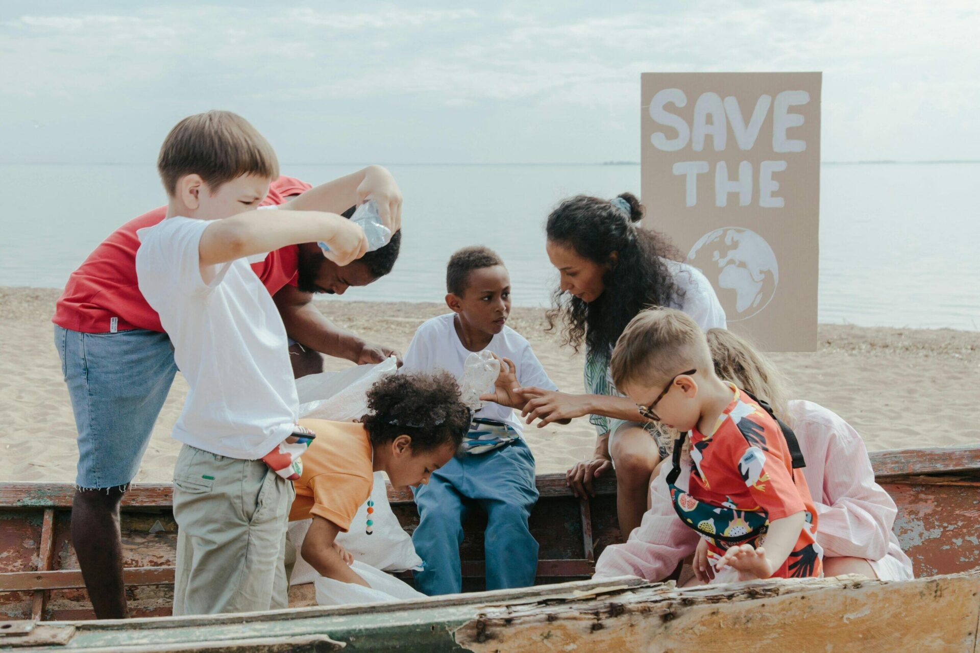 Children and adults collect plastic waste from a boat on a beach, with a "Save the Earth" sign visible.