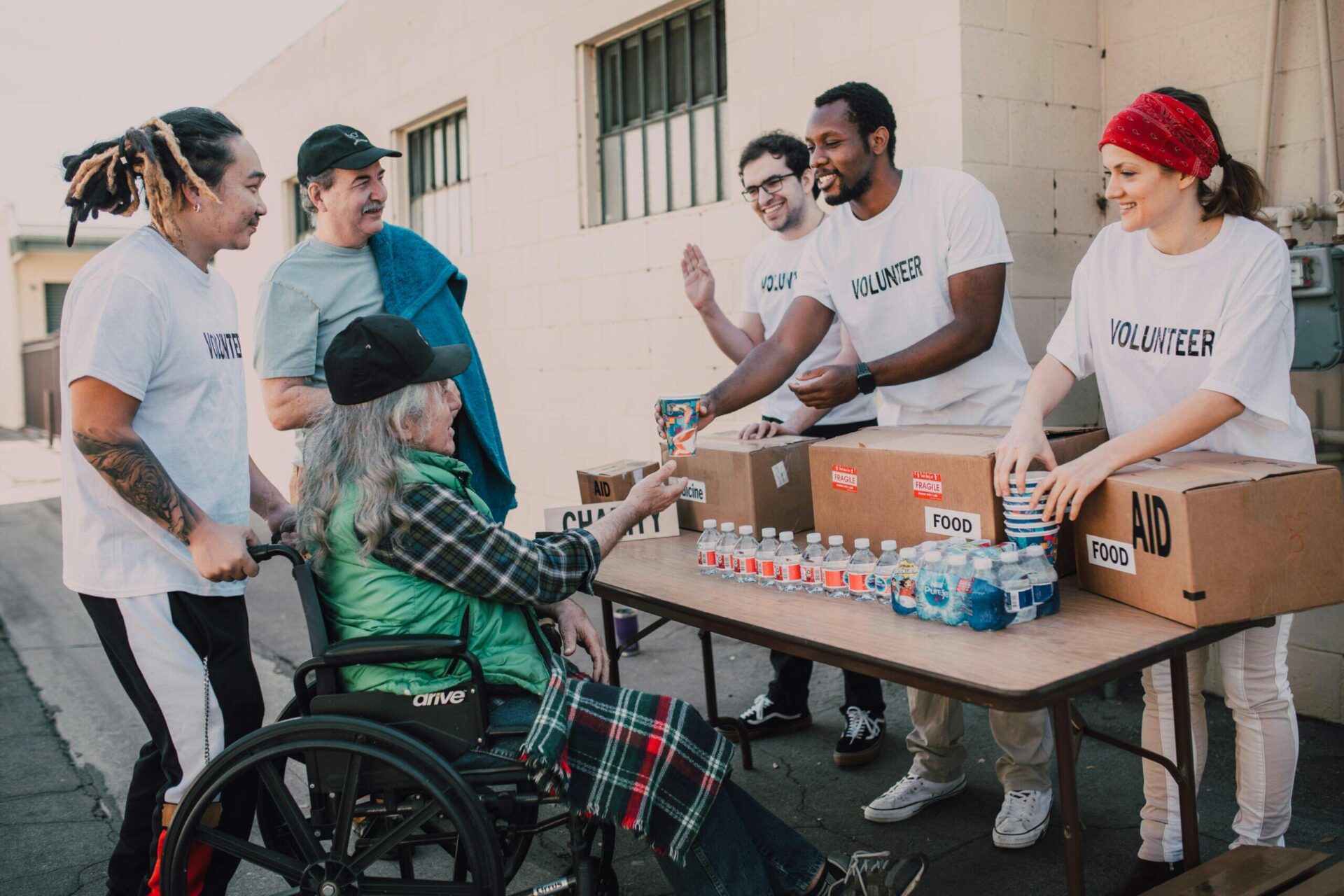 Volunteers distribute food and water to a person in a wheelchair. Boxes labeled "AID" and "FOOD" are visible.