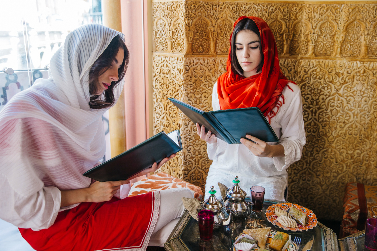 Two women in hijabs reading menus in a cafe with Islamic decor