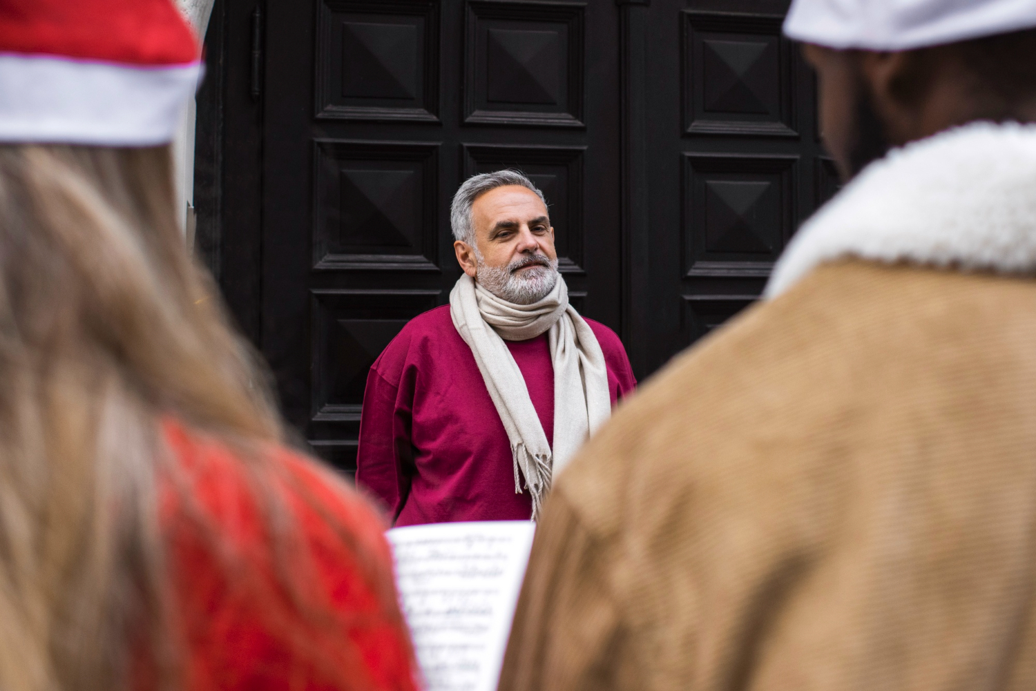 Man with beard and scarf in front of dark door, with people in foreground