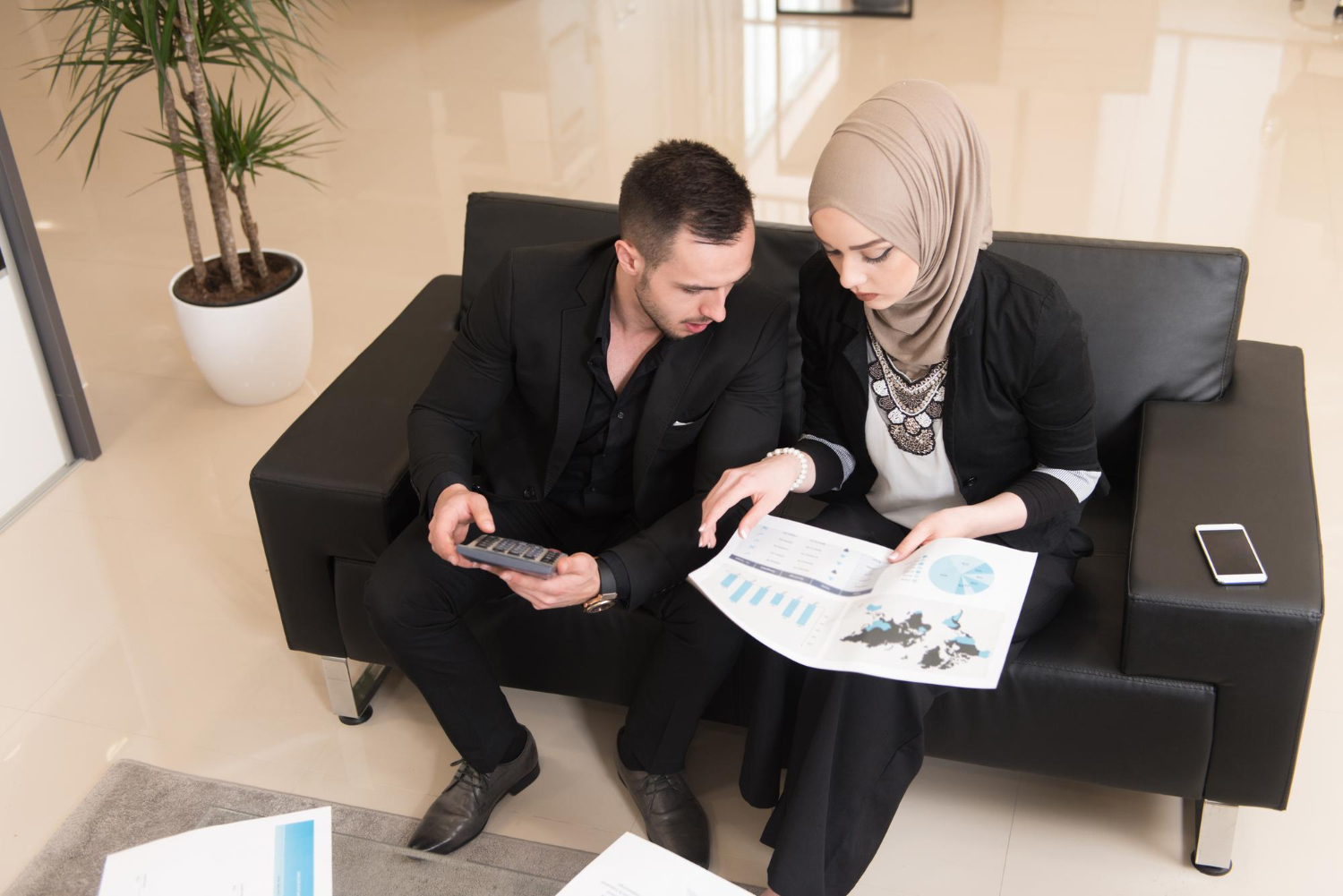 Man and woman in business attire discuss financial documents, with a calculator and smartphone on a couch.